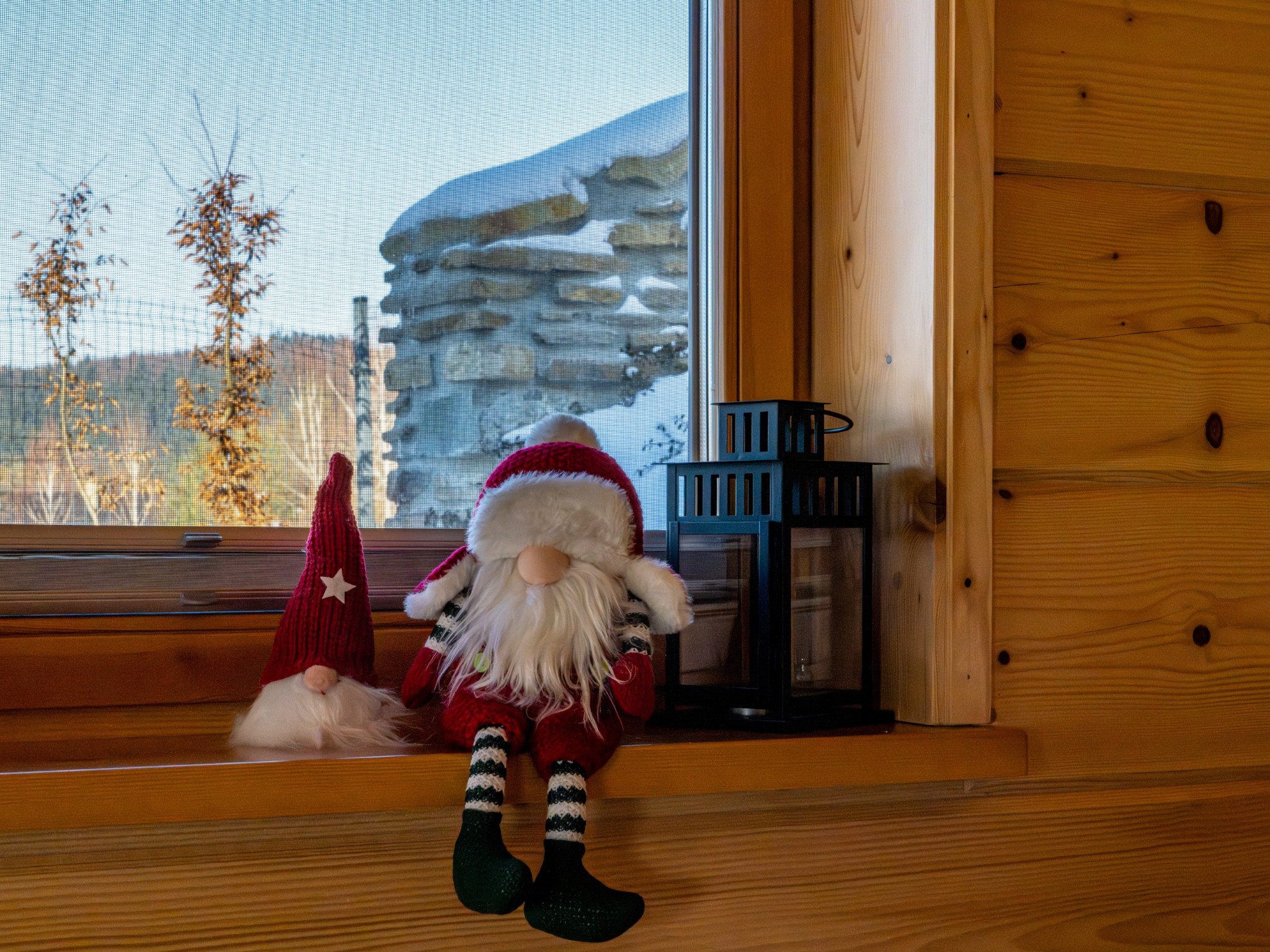 Dwa pluszowe gnomy w czerwonych strojach siedzą na parapecie okna z widokiem na ośnieżony kamienny mur i drzewa. Obok stoi czarny lampion. Drewniane wykończenie wnętrza.