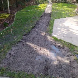 Przygotowany grunt pod trawnik z rolki, widoczna wykopana ścieżka i nawodniona ziemia, obok trawnika leży zwinięty wąż ogrodowy