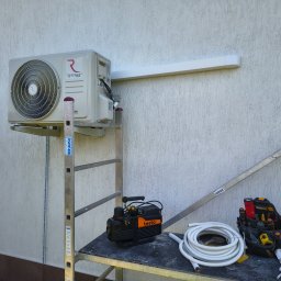 JAKU Jakub Wójcik - Klimatyzator Rotenso zamontowany na zewnątrz budynku, widoczny na rusztowaniu z narzędziami, rurami i torbą narzędziową. Biała ściana z fakturą w tle.