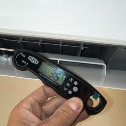 JAKU Jakub Wójcik - Pomiar temperatury klimatyzatora termometrem cyfrowym Refco, trzymanym w dłoni, na tle białej obudowy urządzenia. Widoczny wyświetlacz z wartością 36 stopni Celsjusza.