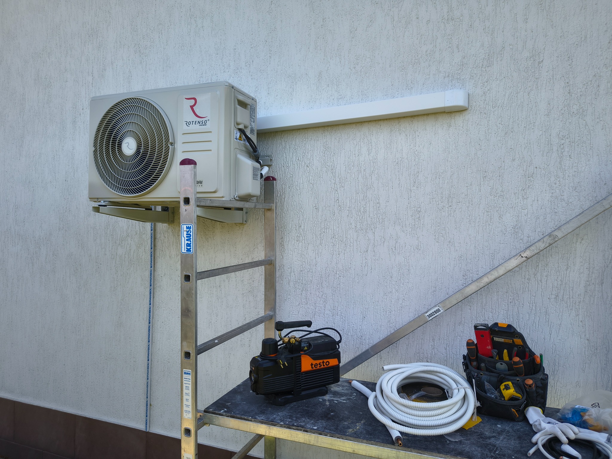 Klimatyzator Rotenso zamontowany na zewnątrz budynku, widoczny na rusztowaniu z narzędziami, rurami i torbą narzędziową. Biała ściana z fakturą w tle.