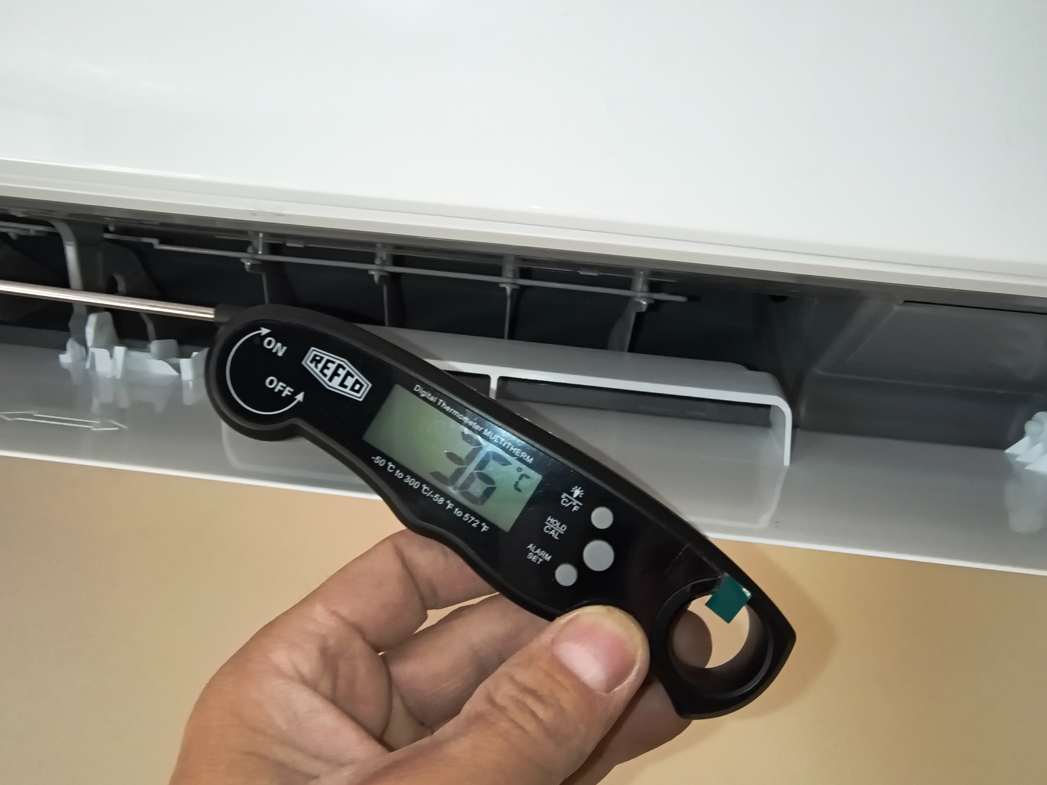Pomiar temperatury klimatyzatora termometrem cyfrowym Refco, trzymanym w dłoni, na tle białej obudowy urządzenia. Widoczny wyświetlacz z wartością 36 stopni Celsjusza.
