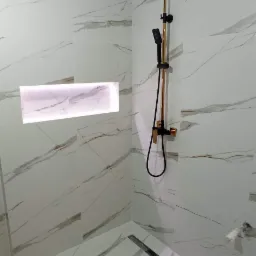 Wykończona kabina prysznicowa z białymi płytkami imitującymi marmur, czarną armaturą z elementami w kolorze szczotkowanego złota oraz podświetlaną wnęką w ścianie.