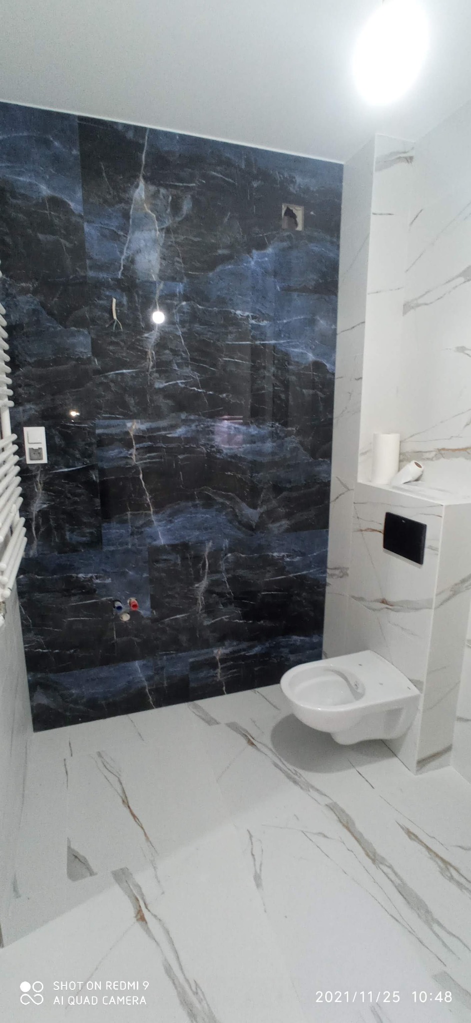 Wykończona łazienka z ciemnymi płytkami imitującymi marmur na ścianie, białą ceramiką sanitarną i grzejnikiem drabinkowym po lewej stronie.