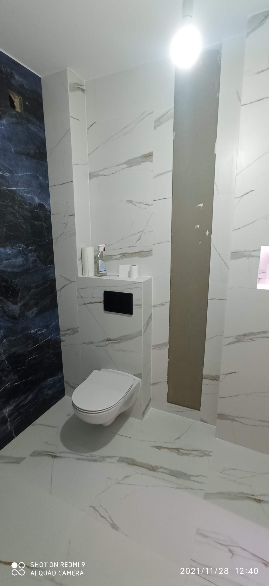 Nowoczesna toaleta podwieszana w łazience wykończonej białymi płytkami imitującymi marmur, z akcentem ciemnych płytek po lewej stronie i widocznym fragmentem niezakończonej obudowy po prawej.