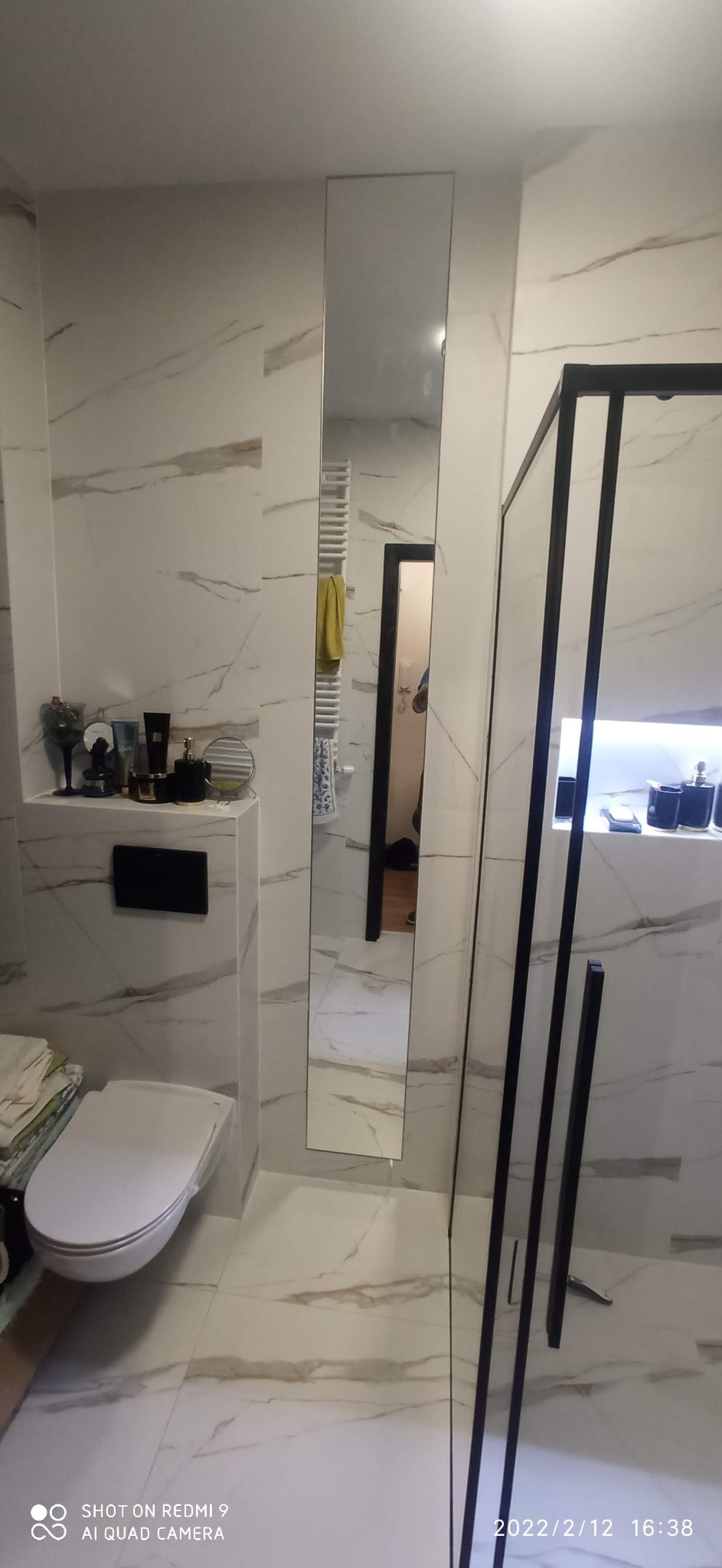 Nowoczesna łazienka z białymi marmurkowymi płytkami, podwieszaną toaletą, wysokim lustrem i kabiną prysznicową z czarnymi ramami.