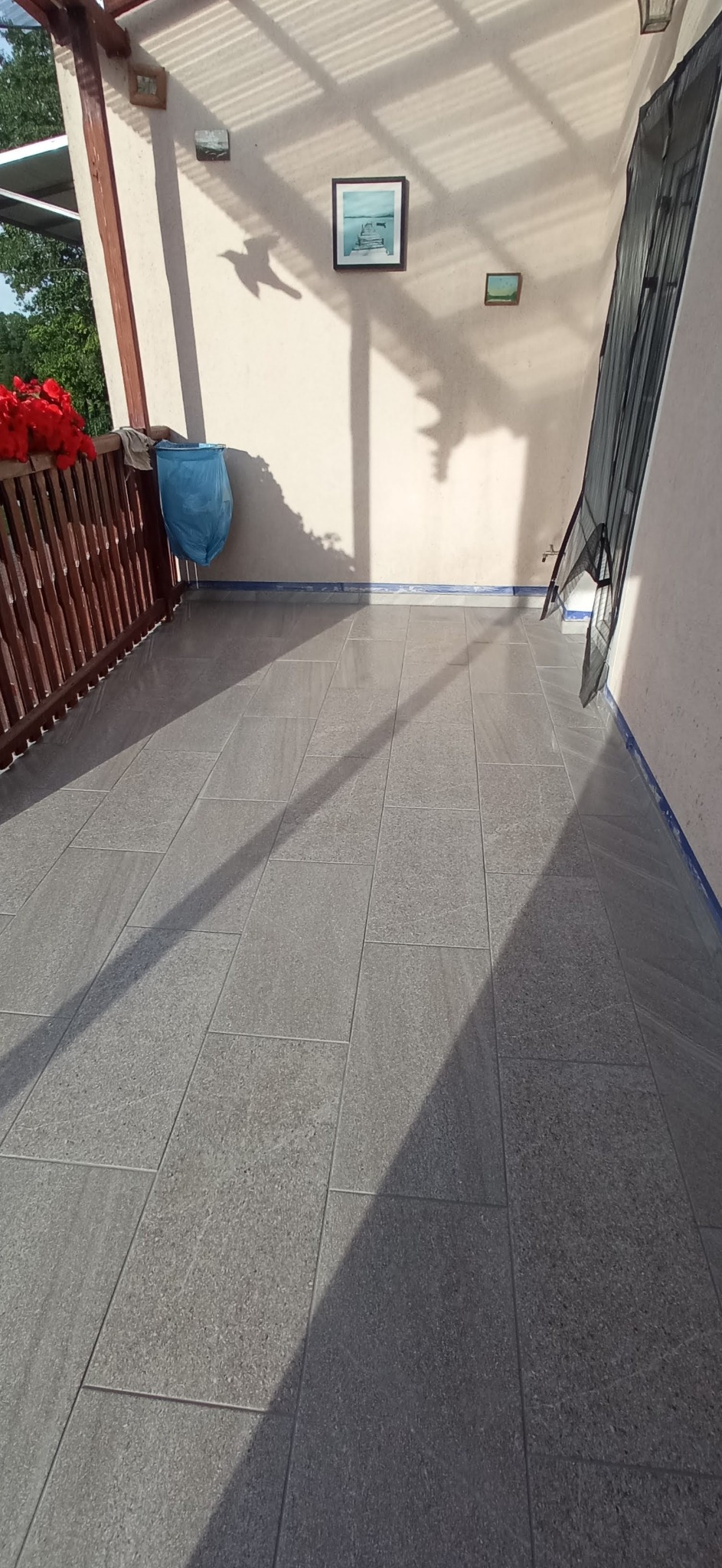 Wyłożony szarymi płytkami taras z drewnianą balustradą, na ścianie wiszą obrazy, widoczne cienie od zadaszenia.