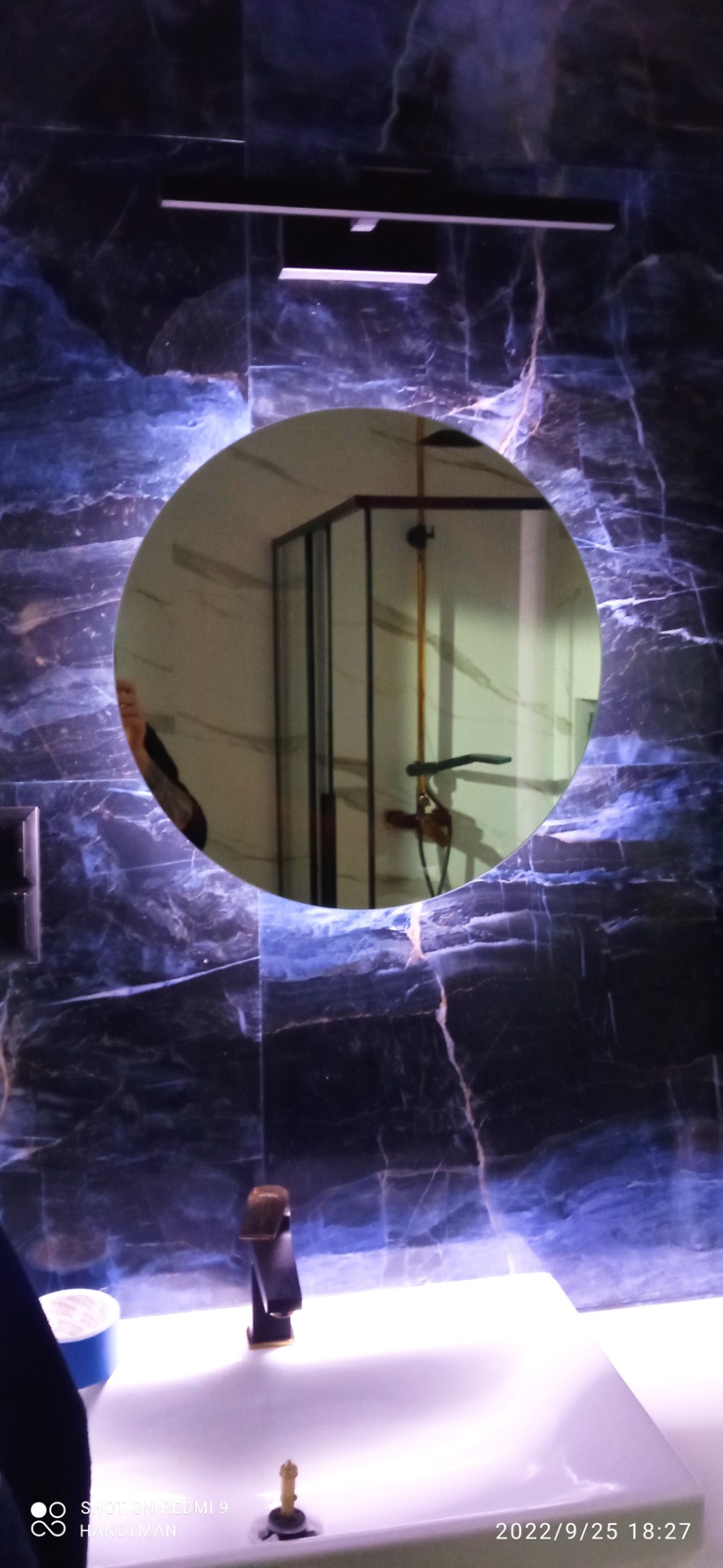 Łazienka z umywalką i baterią w kolorze mosiądzu, ściany wykończone marmurem w odcieniach granatu i błękitu, okrągłe lustro odbijające kabinę prysznicową.