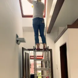 Kobieta na rozstawionej drabinie czyści wewnętrzną stronę okna dachowego w jasnym pomieszczeniu o wysokim suficie z drewnianymi belkami. Widoczny fragment logo firmy na koszulce, oraz osoba w tle.