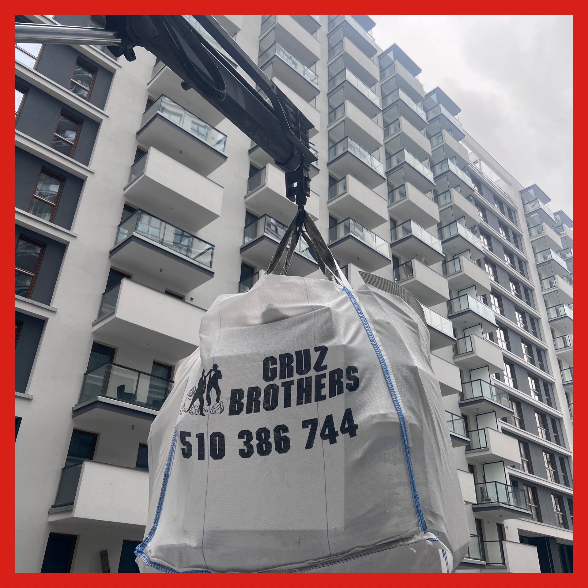 Kran podnosi biały worek Big Bag z napisem 'Gruz BROTHERS' i numerem telefonu na tle nowoczesnego bloku mieszkalnego z balkonami.
