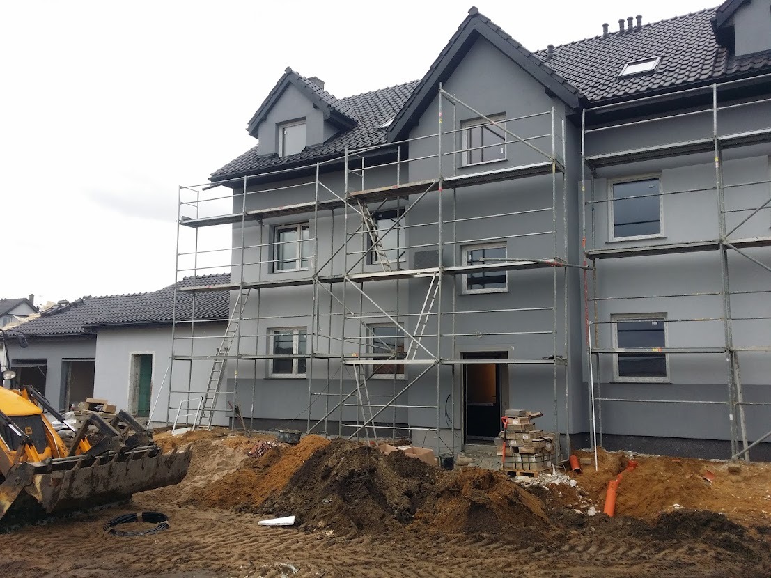 Szary dom w trakcie malowania elewacji, z rusztowaniem, stertami ziemi i koparką w tle, widoczne cegły i rury kanalizacyjne.