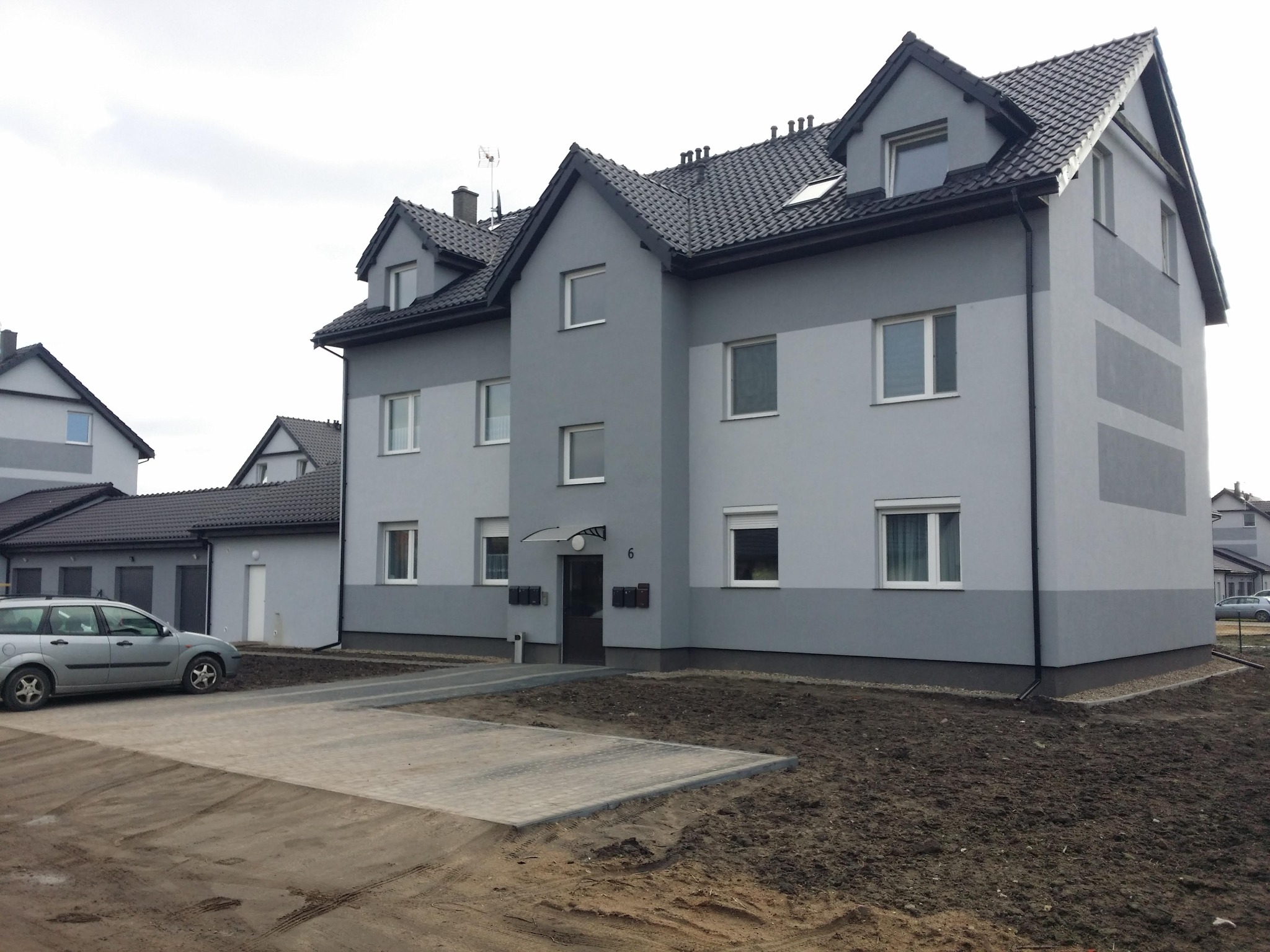 Nowoczesny, dwukolorowy budynek mieszkalny z ciemną dachówką i numerem 6 nad wejściem, otoczony świeżo przygotowanym terenem pod trawnik i parkingiem z kostki.