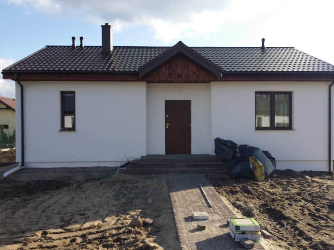 Nowoczesny dom jednorodzinny z białą elewacją, brązowym dachem z dachówki i brukowanym chodnikiem w trakcie układania, widoczne worki z odpadami budowlanymi.