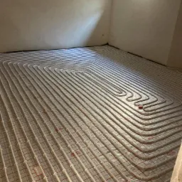 Układanie rur ogrzewania podłogowego na folii z siatką w surowym wnętrzu, widoczne okno w tle.