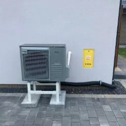 Szara jednostka zewnętrzna urządzenia grzewczego, zamontowana na białym stelażu przy ścianie domu w Oławie, obok skrzynki gazowej.