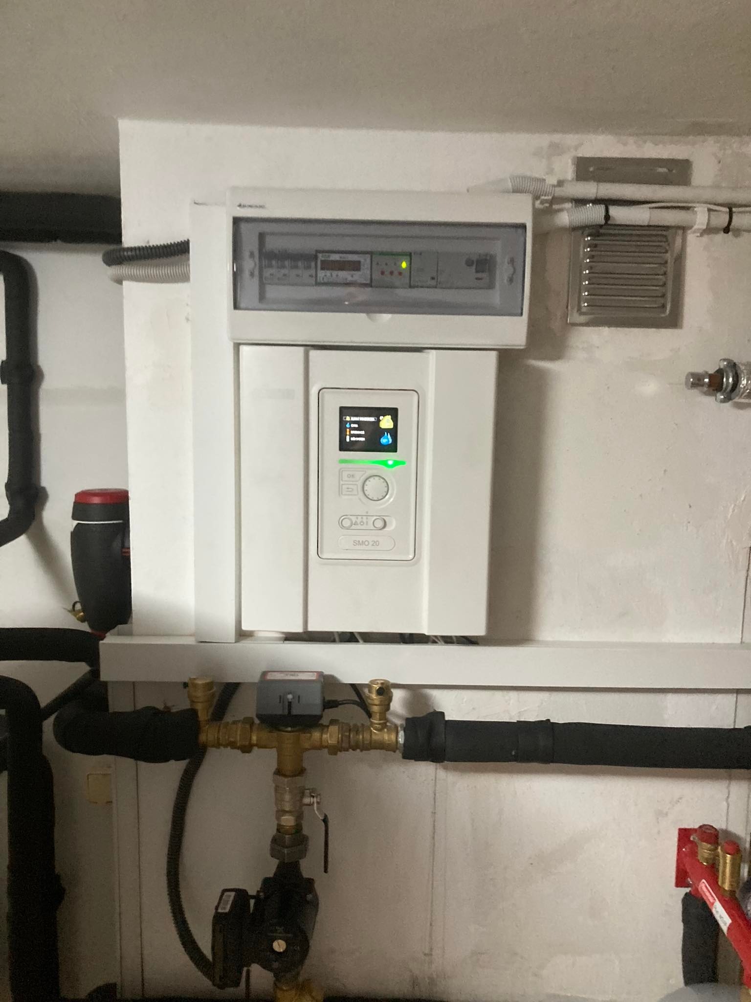 Biała centrala grzewcza z wyświetlaczem i panelem sterowania, zamontowana na ścianie nad instalacją hydrauliczną z czarnymi rurami i pompą obiegową.
