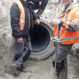 Trzech pracowników w kaskach i kamizelkach odblaskowych w Gdańsku, podczas instalacji rury kanalizacyjnej, jeden z nich używa urządzenia do pomiaru zagęszczenia gruntu Terratest 5000.