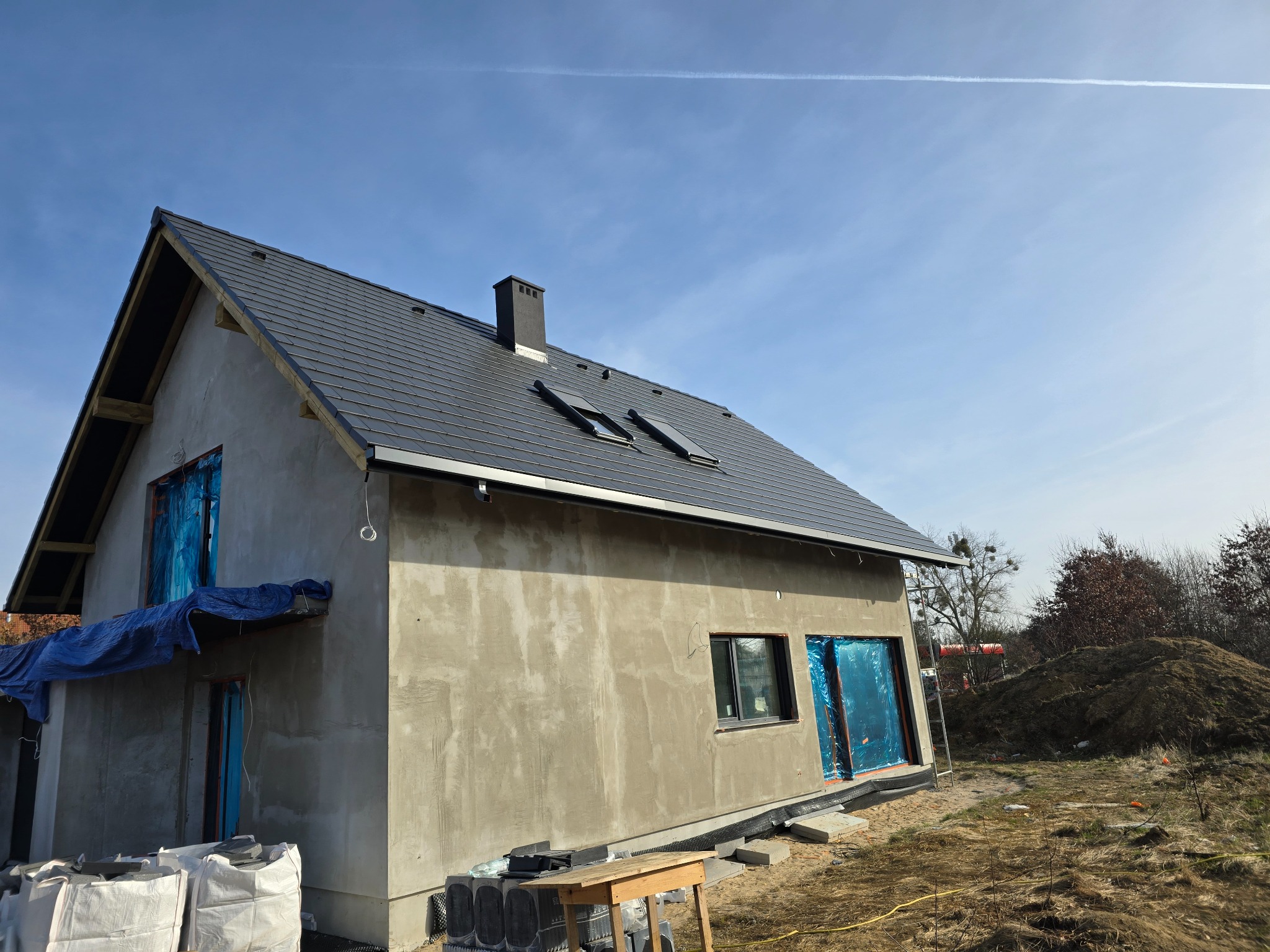 Surowy dom jednorodzinny z ciemną dachówką i oknami połaciowymi, elewacja w trakcie wykończenia, okna i drzwi zabezpieczone folią, widoczne worki z materiałami budowlanymi i drewniany stół.