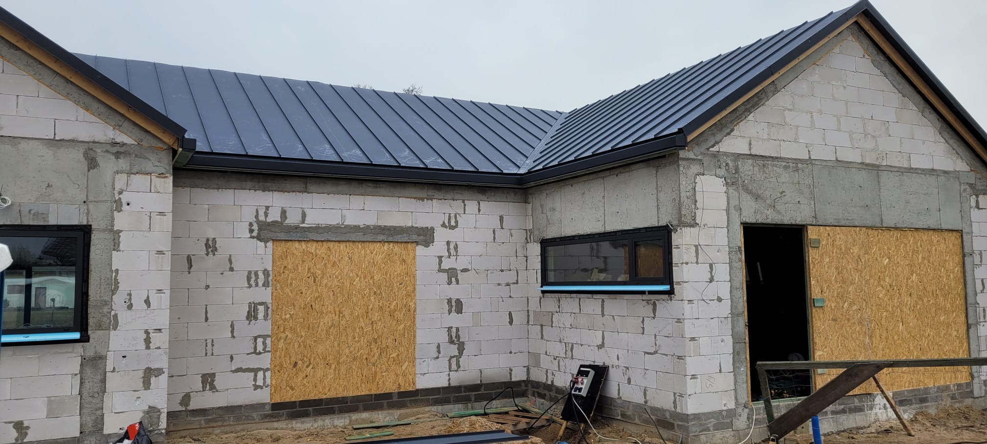 Surowy stan budynku z szarego bloczka, z zamontowaną, ciemną blachą na dachu i czarnymi rynnami, okna zabezpieczone folią, otwory okienne i drzwiowe zabite płytami OSB.