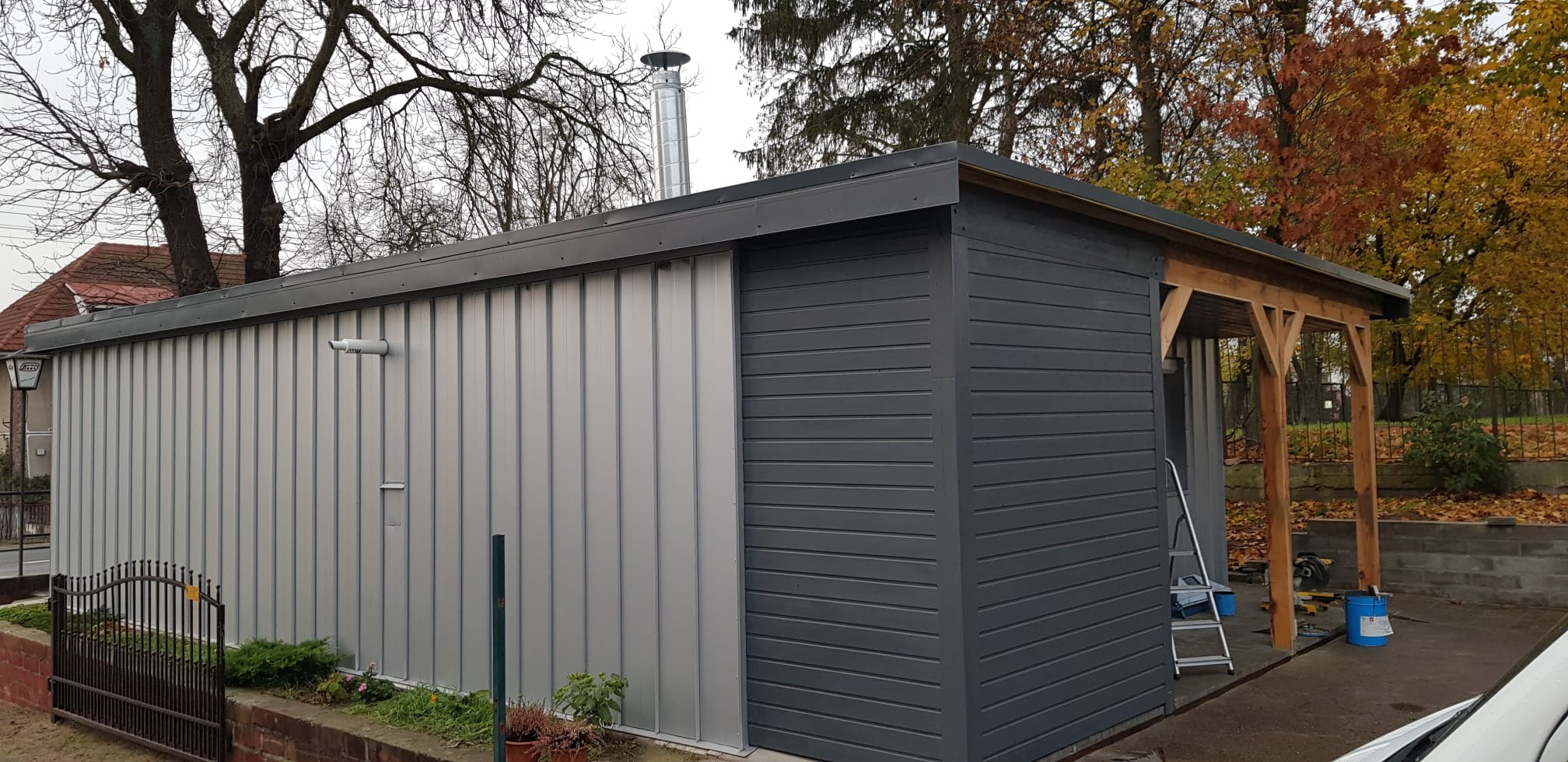 Nowoczesna konstrukcja z blachy trapezowej i szarej boazerii, z drewnianą konstrukcją wnęki, widoczny komin stalowy i drabina oparta o ścianę.