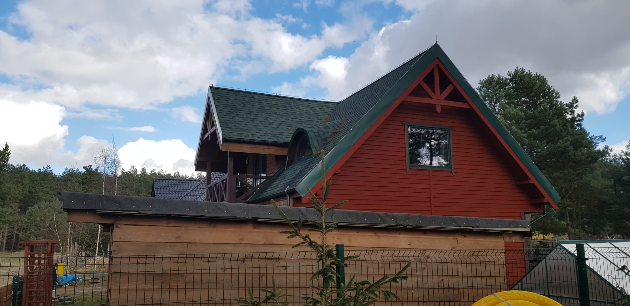 Czerwony dom z zielonym dachem bitumicznym, widok zza ogrodzenia z siatki, na tle lasu i błękitnego nieba z chmurami.