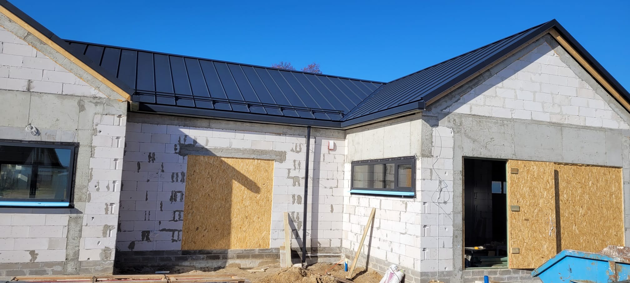 Budynek w trakcie budowy z zamontowanym, czarnym dachem z blachy na rąbek stojący i orynnowaniem, okna zabezpieczone folią, otwory okienne i drzwiowe zabite płytami OSB.