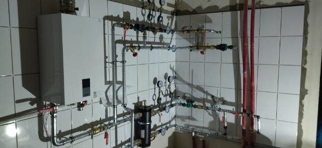 Biały piec gazowy kondensacyjny wiszący na ścianie z białych płytek, z widoczną rozbudowaną instalacją hydrauliczną z licznymi zaworami i manometrami.