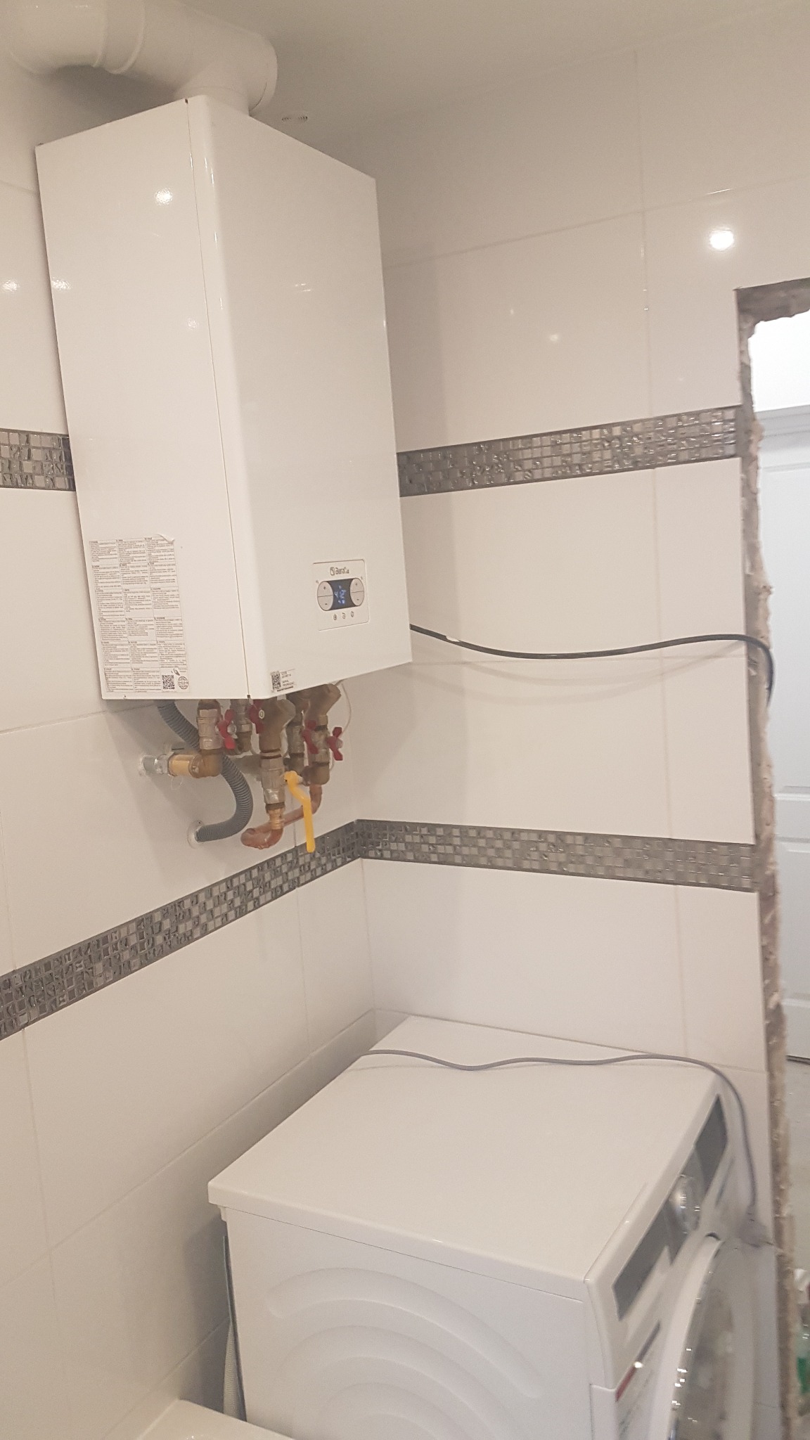 Biały piec gazowy marki Beretta zamontowany na ścianie z białych płytek z dekoracyjnym pasem mozaiki, widoczne przyłącza hydrauliczne i gazowe, poniżej pralka.