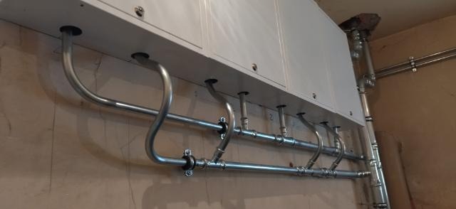 Biały piec gazowy zamontowany na ścianie z widocznymi rurami instalacji gazowej, z charakterystycznymi kolankami i mocowaniami, prowadzącymi do otworów w obudowie pieca.