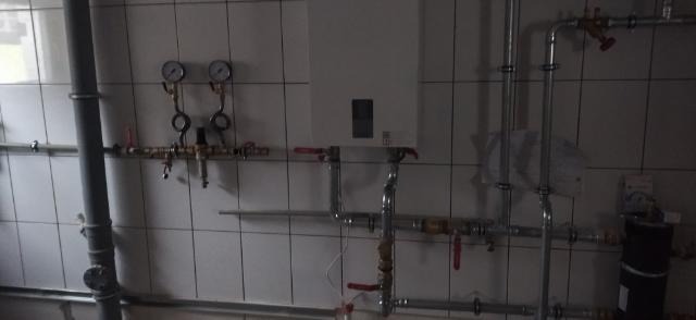 Biały piec gazowy zamontowany na ścianie z białych płytek, widoczne rury, zawory i manometry, instalacja w trakcie podłączania, widoczne przewody elektryczne.