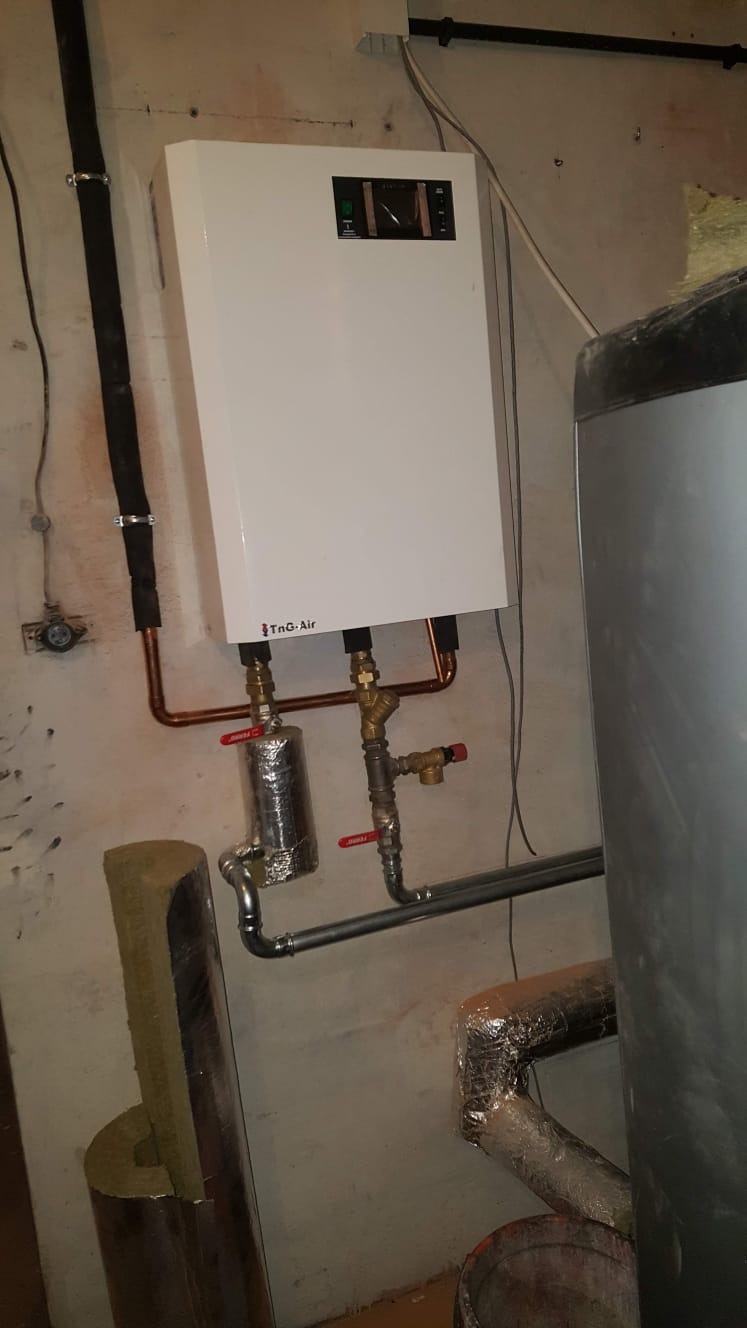Biały piec grzewczy marki TnG-Air zamontowany na szarej, surowej ścianie piwnicy, widoczne miedziane rury, zawory z czerwonymi rączkami, izolowane rury i fragment zasobnika wody.