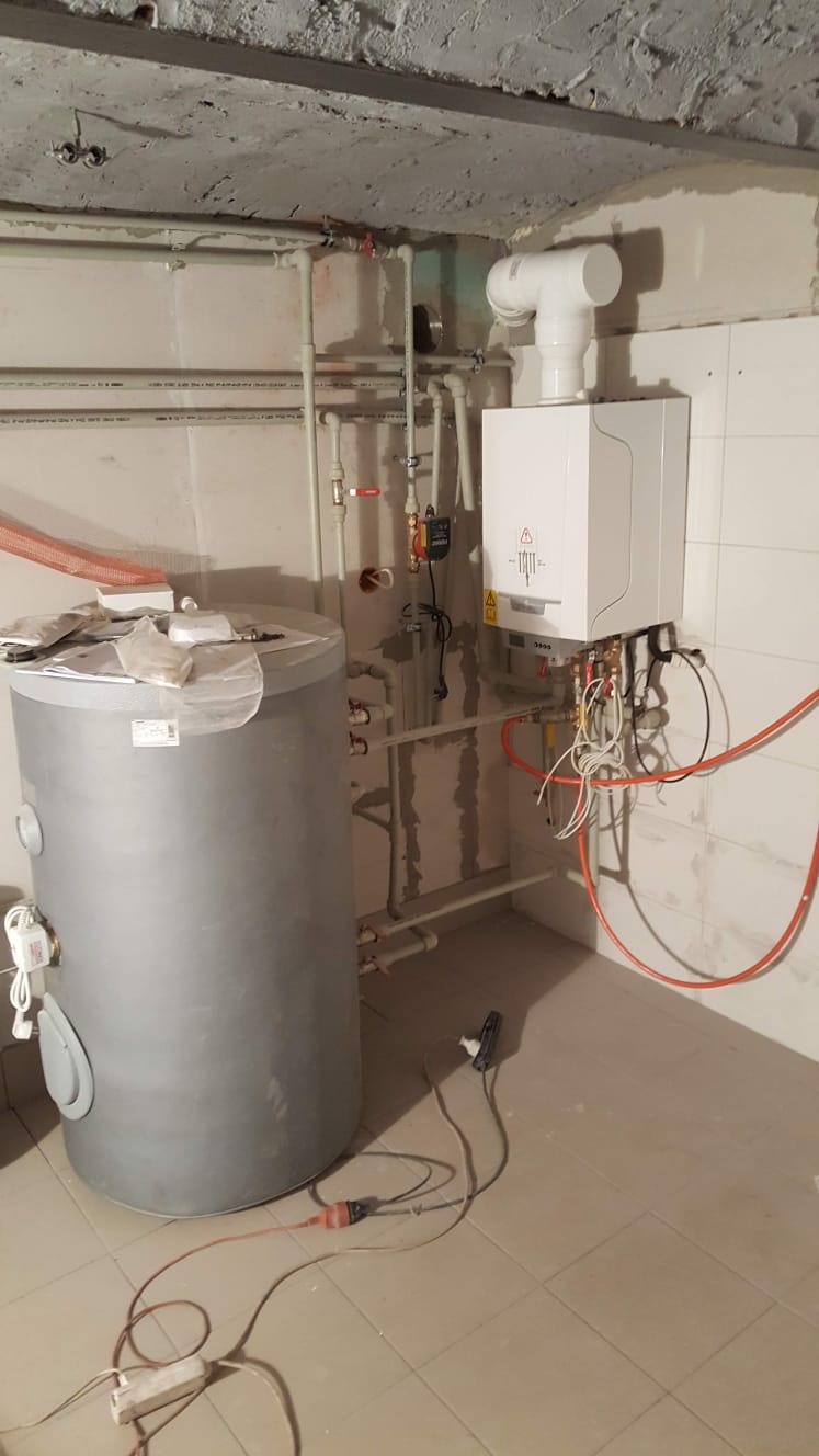 Nowy, biały piec gazowy kondensacyjny zamontowany na ścianie w surowym pomieszczeniu z widocznymi rurami, zbiornikiem na wodę i przewodami elektrycznymi, z surowym betonowym sufitem...