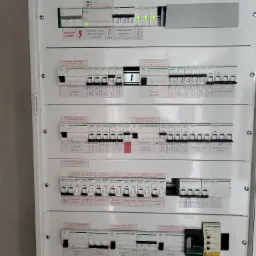 Rozdzielnica elektryczna z licznymi zabezpieczeniami, wyłącznikami różnicowoprądowymi i opisami obwodów, widoczne naklejki informacyjne o konieczności testowania wyłącznika RCCB.