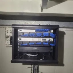 Szafa rack z zamontowanym rejestratorem Dahua i patch panelem, uporządkowane niebieskie kable sieciowe, instalacja w pomieszczeniu technicznym z widocznymi elementami konstrukcyjnymi sufitu.