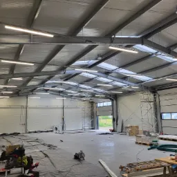 Wnętrze hali produkcyjnej w trakcie prac instalacyjnych oświetlenia, widoczne lampy liniowe pod sufitem, rusztowania i materiały budowlane na podłodze zabezpieczonej folią.