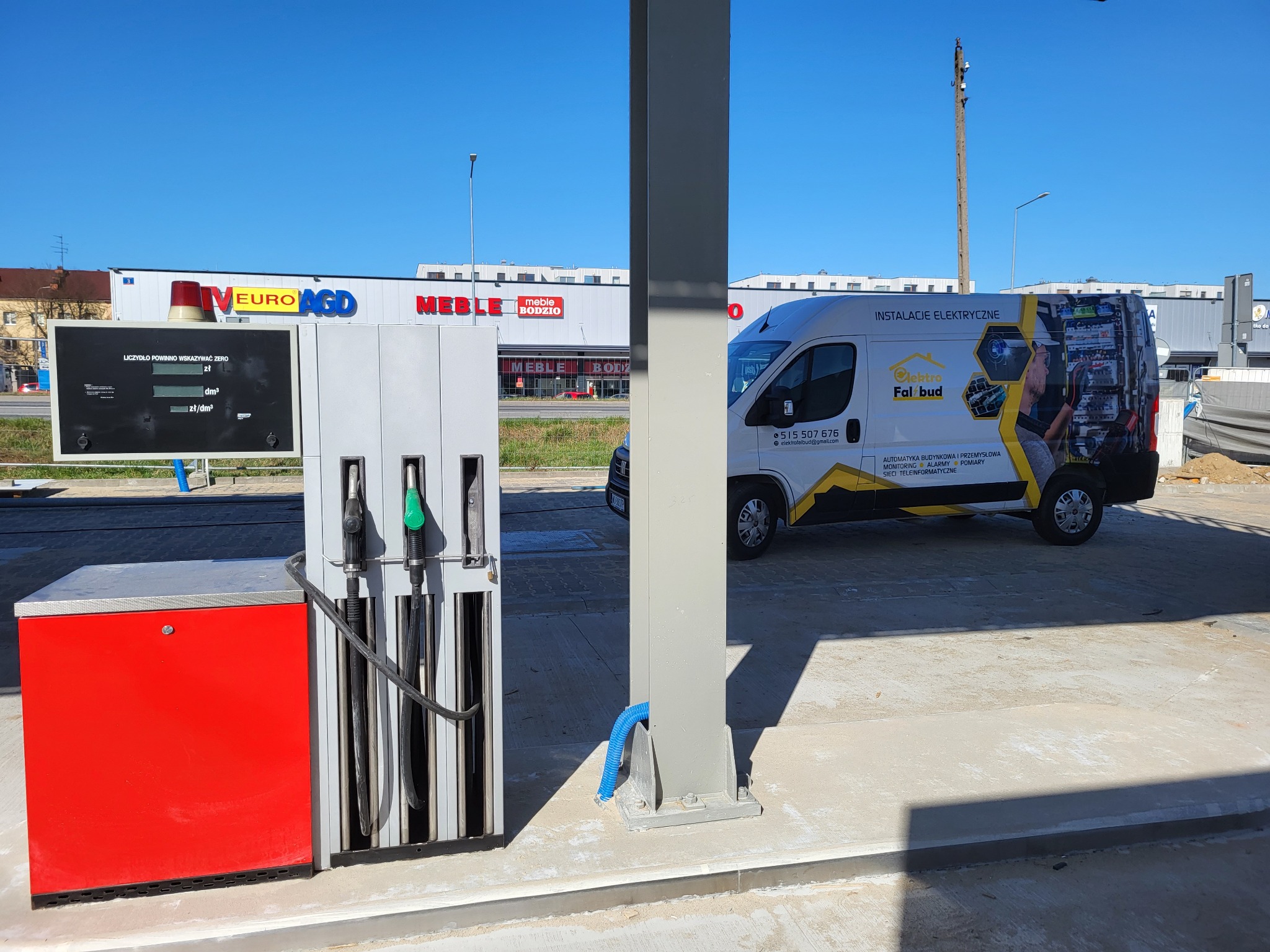 Furgonetka firmy Elektro-Falbud z reklamą usług elektrycznych zaparkowana na stacji benzynowej w słoneczny dzień, widoczny dystrybutor paliwa i budynki sklepów w tle.