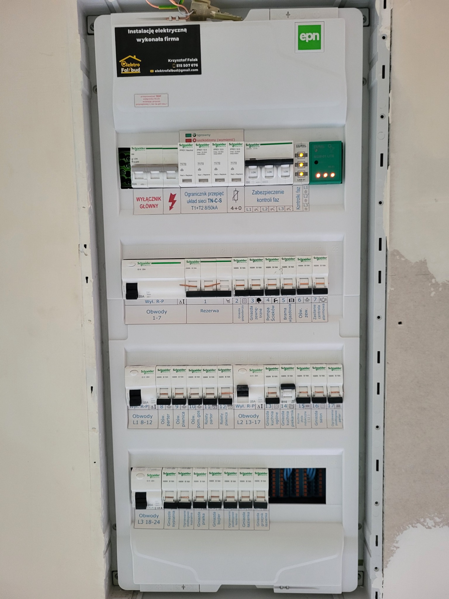 Rozdzielnica elektryczna w białej obudowie, z bezpiecznikami i opisami obwodów, zainstalowana w nowym budynku z widocznymi śladami po pracach wykończeniowych, z naklejką informacyjną firmy...