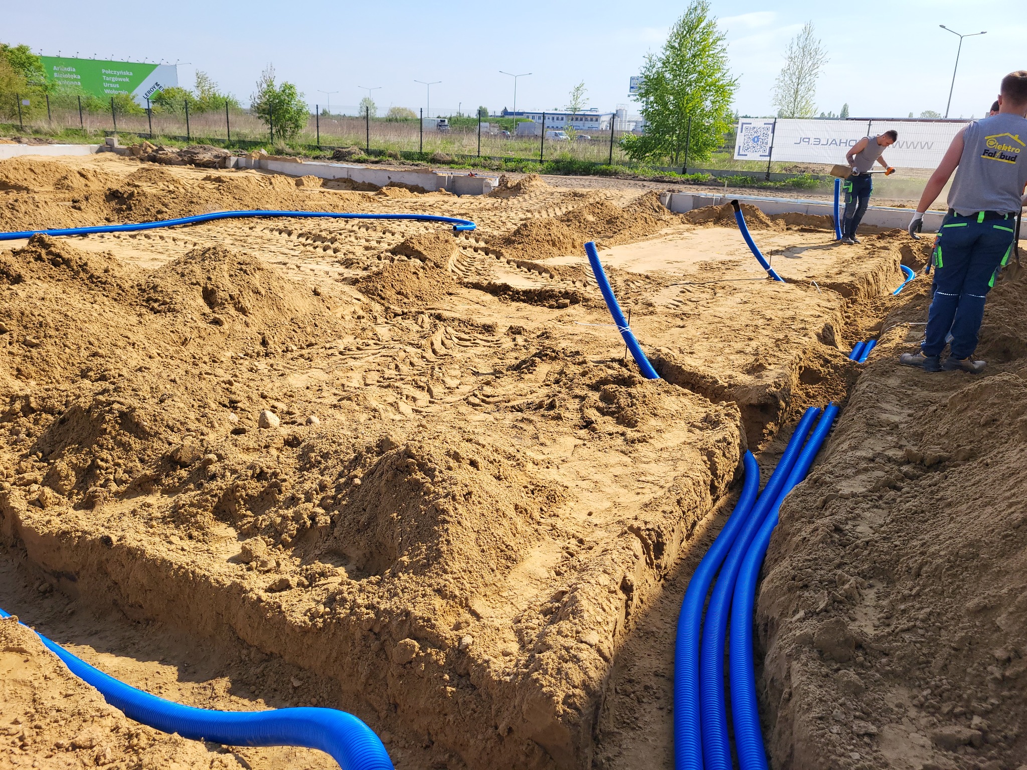 Wykop pod fundamenty z ułożonymi niebieskimi rurami karbowanymi; w tle pracownicy z łopatami i reklama zewnętrzna.