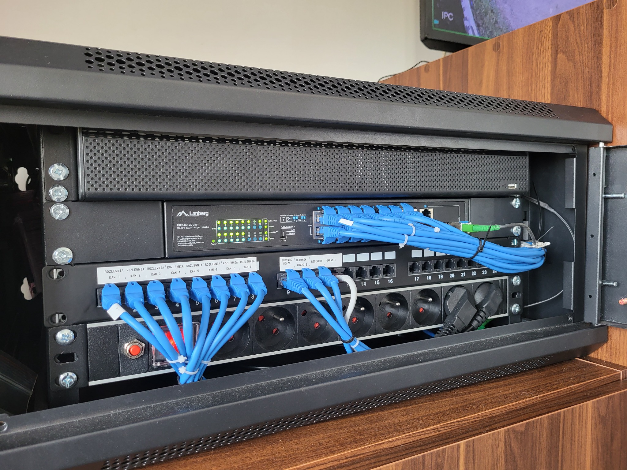 Szafa rack z zainstalowanym switchem Lanberg, patch panelem i listwą zasilającą, okablowanie strukturalne z niebieskimi kablami zakończonymi wtykami RJ45, widoczne porty ethernet i gniazda zasilające.