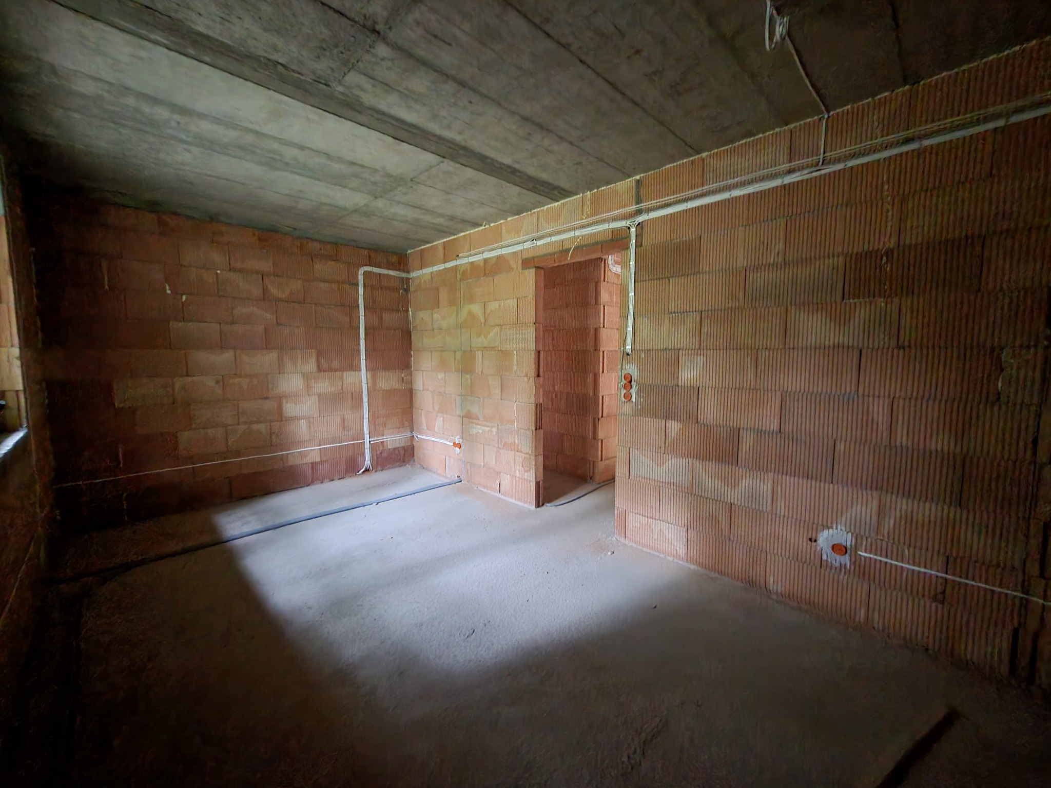 Surowy stan wnętrza z widocznymi przewodami elektrycznymi poprowadzonymi po ścianach z cegły, sufit betonowy, brak wykończenia.