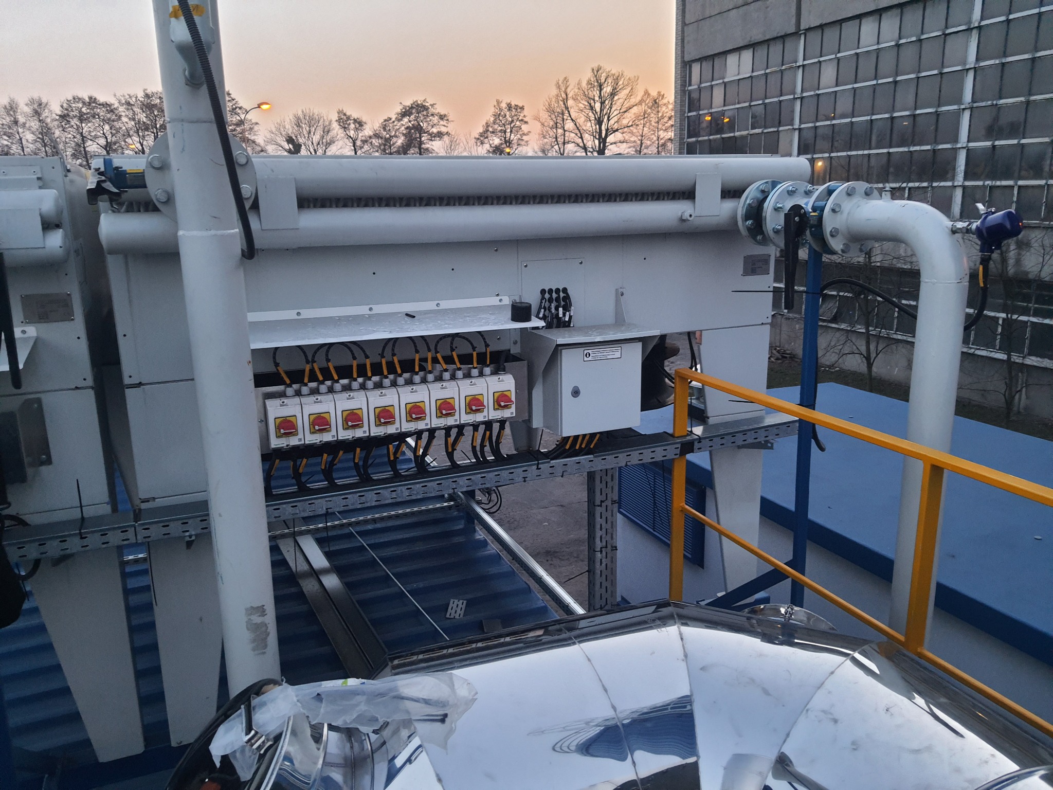 Biała przemysłowa instalacja z przełącznikami i rurami na tle budynku i wieczornego nieba.