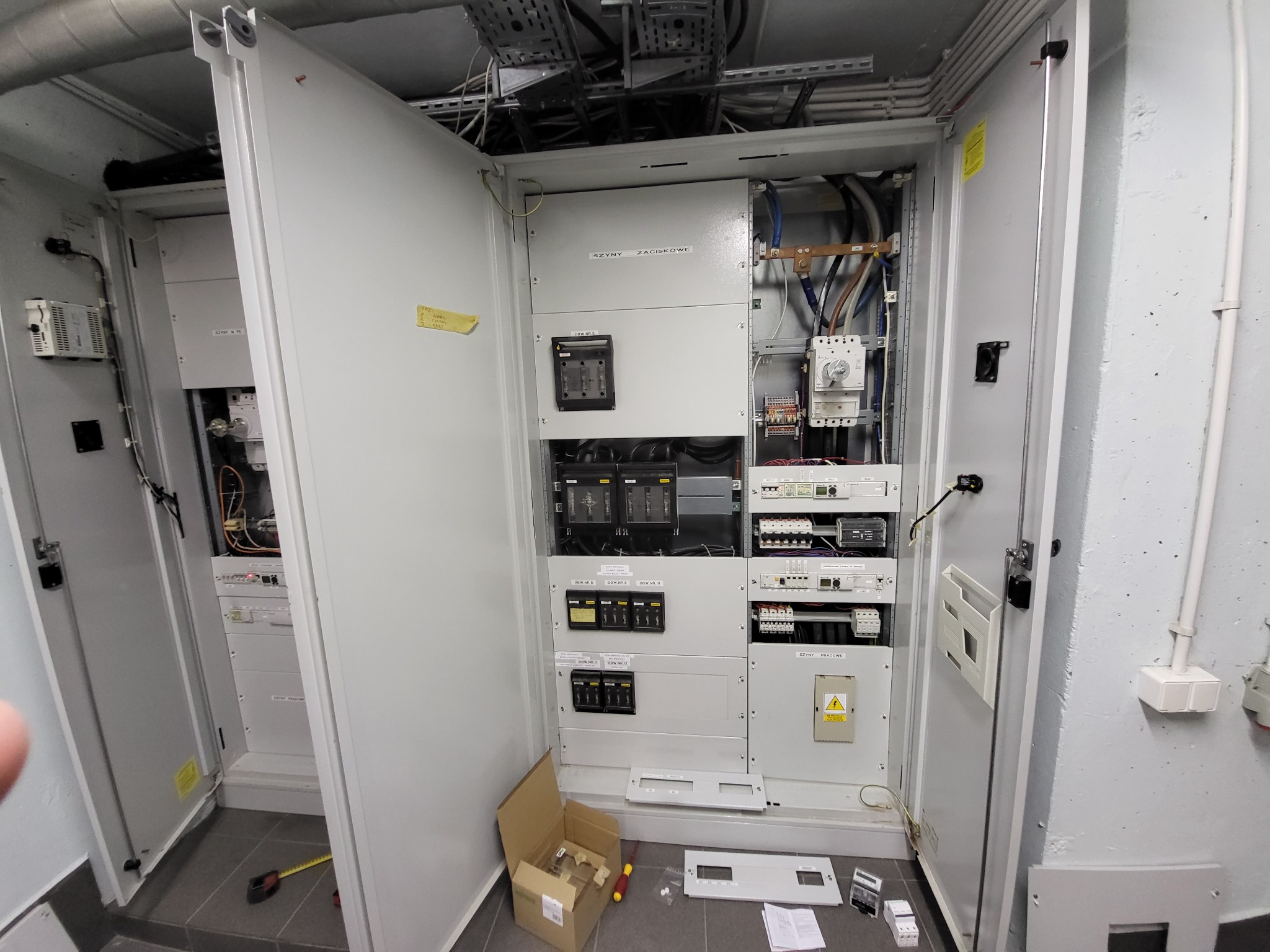 Otwarta szafa elektryczna z widocznymi zabezpieczeniami, przewodami, skrzynką narzędziową i elementami montażowymi na podłodze, sugerującymi etap prac instalacyjnych.