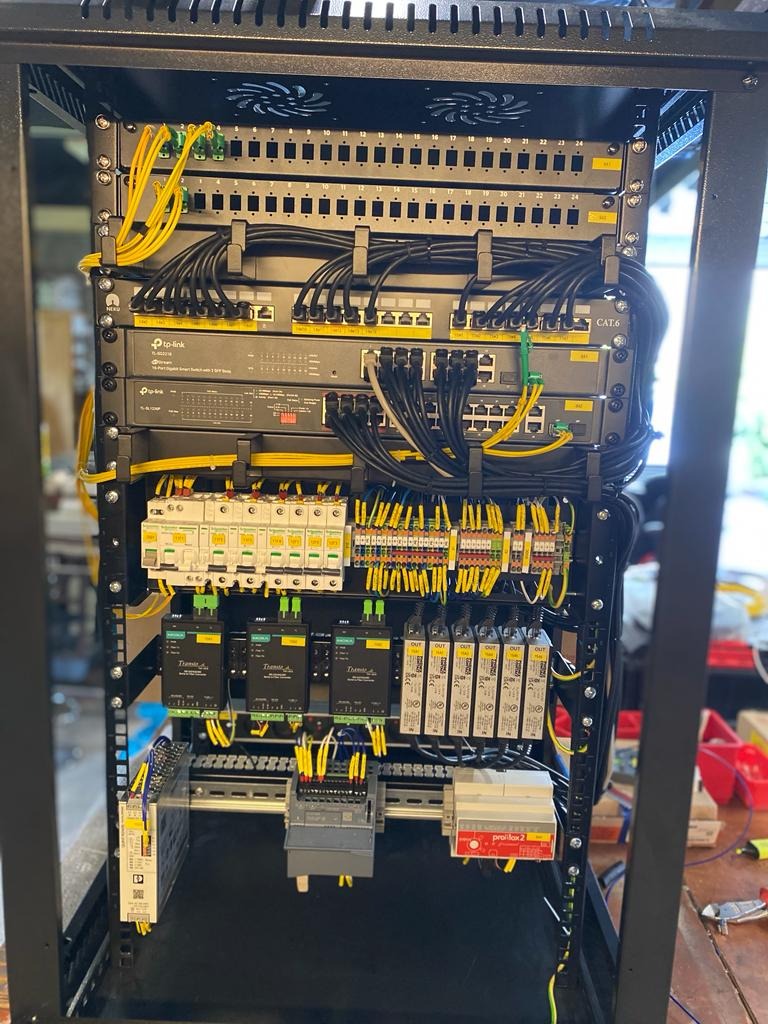 Rozdzielnia elektryczna w metalowej obudowie, z widocznymi zabezpieczeniami, modułami sterującymi, przełącznikami sieciowymi TP-Link oraz licznymi kablami w kolorze żółtym i czarnym, starannie...