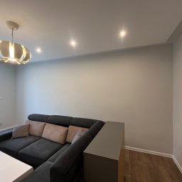 AD-REM FIRMA USŁUGOWA ADAM GRACZYK - Jasne wnętrze po malowaniu: szare ściany, drewniana podłoga, nowoczesna lampa i narożnik z poduszkami w odcieniach beżu. Widoczne listwy przypodłogowe.