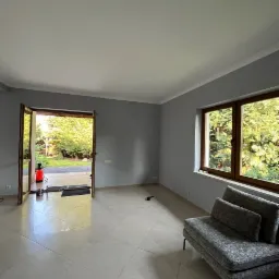 Wnętrze po malowaniu ścian na szaro, widok na okno z zielenią i częściowo widoczny szary narożnik. Podłoga wyłożona jasnymi płytkami. Otwarty balkon z widokiem na ogród.