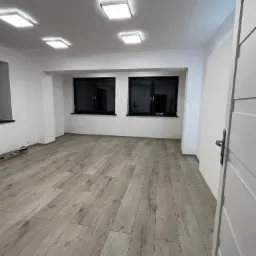 Jasne panele podłogowe ułożone w pustym, odnowionym pokoju z białymi ścianami, czarnymi oknami i otwartymi, białymi drzwiami. Widoczne narzędzia malarskie.