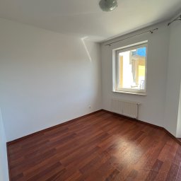 AD-REM FIRMA USŁUGOWA ADAM GRACZYK - Puste, jasne wnętrze po remoncie: ciemne panele podłogowe, białe ściany, okno z grzejnikiem. Widoczny fragment karnisza i lampa sufitowa.