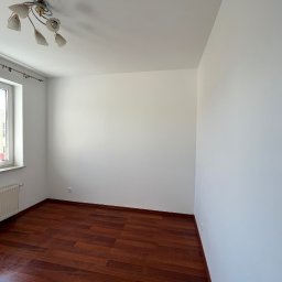 AD-REM FIRMA USŁUGOWA ADAM GRACZYK - Pusty pokój po remoncie: świeżo pomalowane białe ściany, ciemna drewniana podłoga, okno z grzejnikiem i lampa sufitowa. Widok na narożnik pokoju.