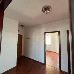 AD-REM FIRMA USŁUGOWA ADAM GRACZYK - Korytarz po remoncie: świeżo pomalowane ściany w odcieniach bieli, ciemne drewniane panele podłogowe, widoczne drzwi wejściowe i przejście do innego pomieszczenia.