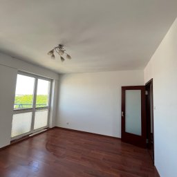 AD-REM FIRMA USŁUGOWA ADAM GRACZYK - Pusty pokój z drewnianą podłogą, białymi ścianami i oknem z żaluzjami. Widoczny otwarty, ciemny dąb drzwi. Lampa sufitowa w stylu retro. Jasne, naturalne światło.